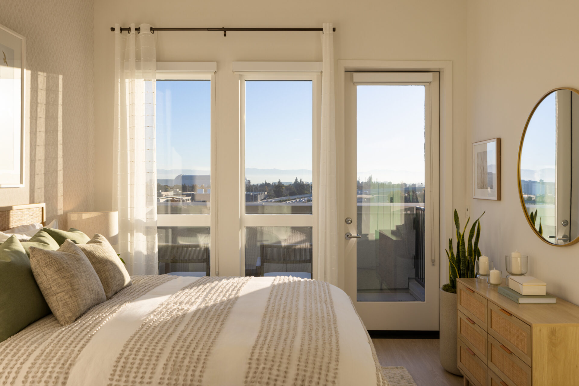 Mountain View, CA, Luxury Apartments Near Levis Stadium - Avelle - Sunlit Bedroom With Large Windows, Balcony Door, Neutral Bedding, Light Wood Dresser, And Round Wall Mirror.